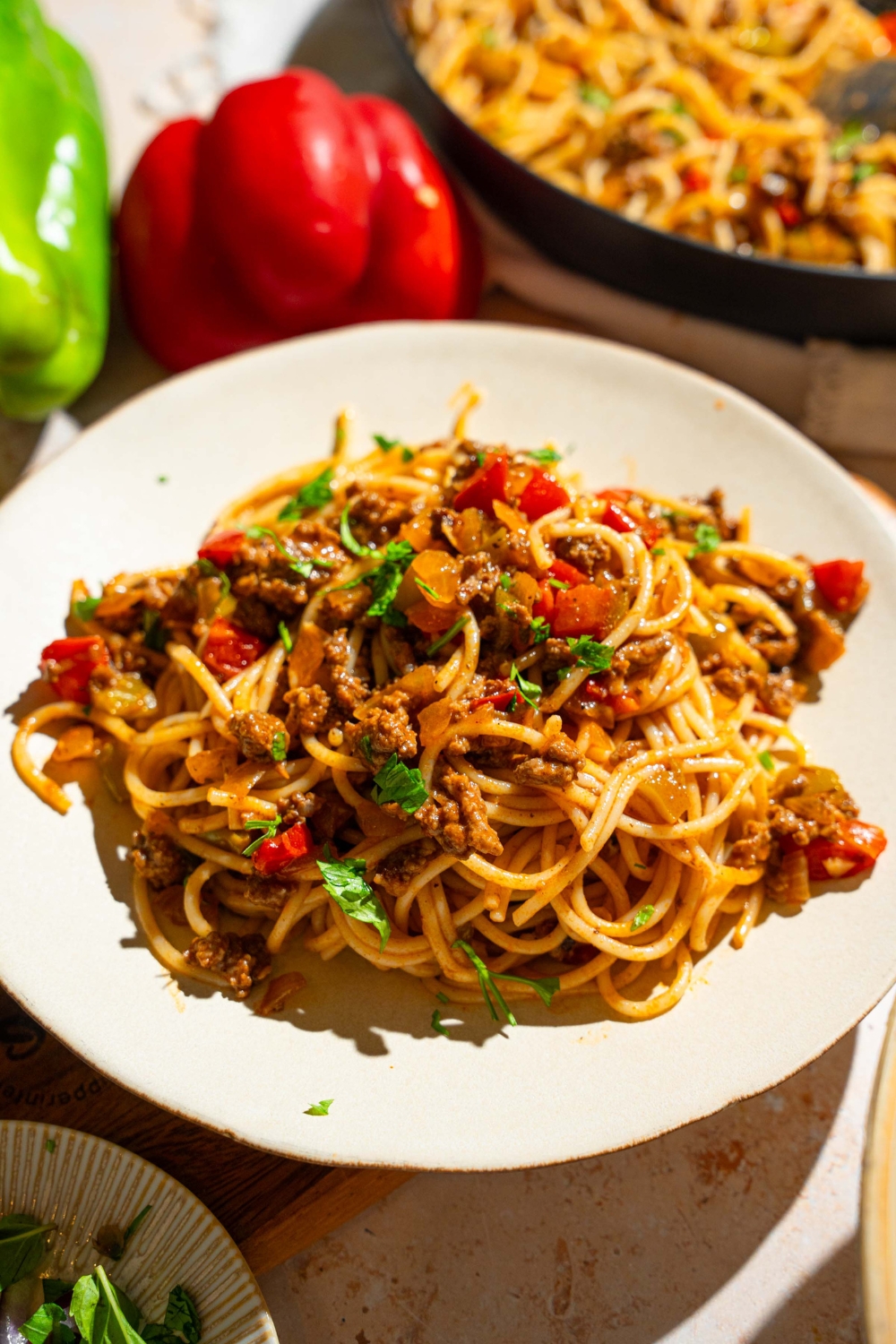 A white plate with dirty spaghetti tossed with diced peppers, celery, onion, ground beef, and Italian sausage garnished with fresh parsley. The plate is on a tan counter with a skillet of spaghetti and peppers.