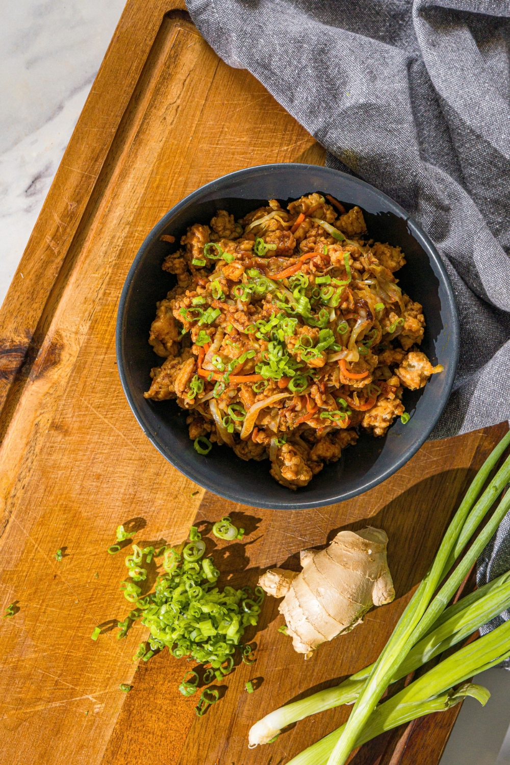 A navy bowl with an egg roll in a bowl, made with ground chicken and sautéed cabbage and carrots tossed in an egg roll sauce, all garnished with sliced green onions. The bowl is on a wooden board with sliced green onion and ginger.
