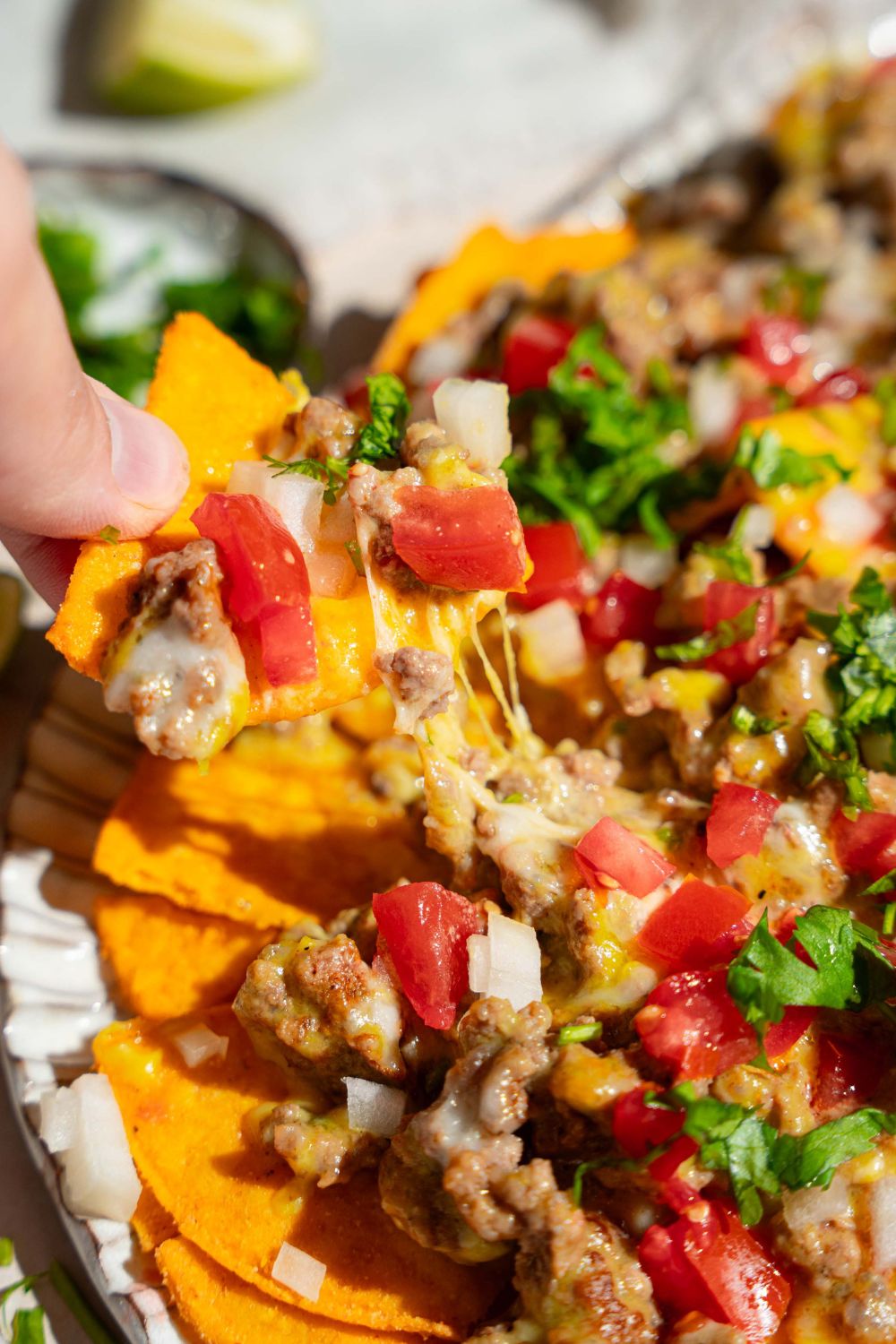 An oval platter with sliding sheet pan nachos topped with shredded cheese, ground beef, diced tomatoes and onions, and fresh parsley. A nacho is being pulled from the platter. The platter is on a tan counter.