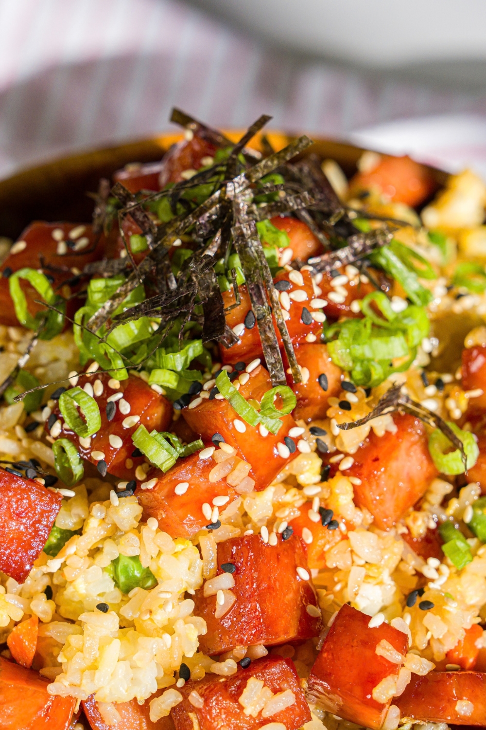 A spam musubi rice bowl garnished with seaweed, green onion, and sesame seeds. The bowl is on a wooden board with small bowls of garnishes.
