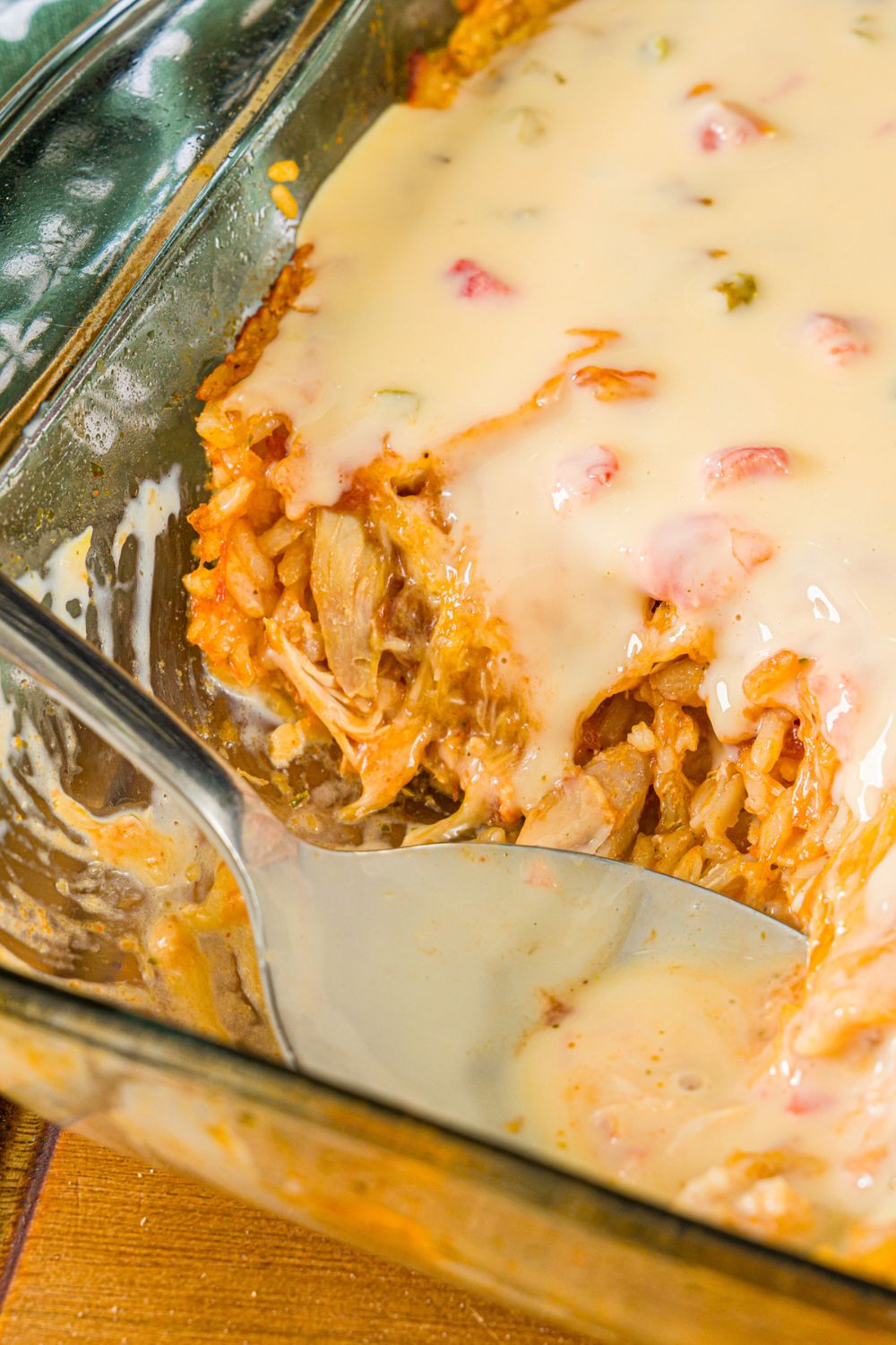A baking dish with swim queso chicken and rice. A spoon is taking a scoop from the dish. The dish is on a wooden board.