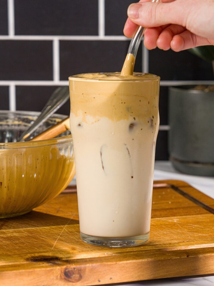 A glass with whipped honey iced coffee with milk. There is a spoon stirring the coffee in the glass. The glass is on a wooden board with a green cloth napkin.