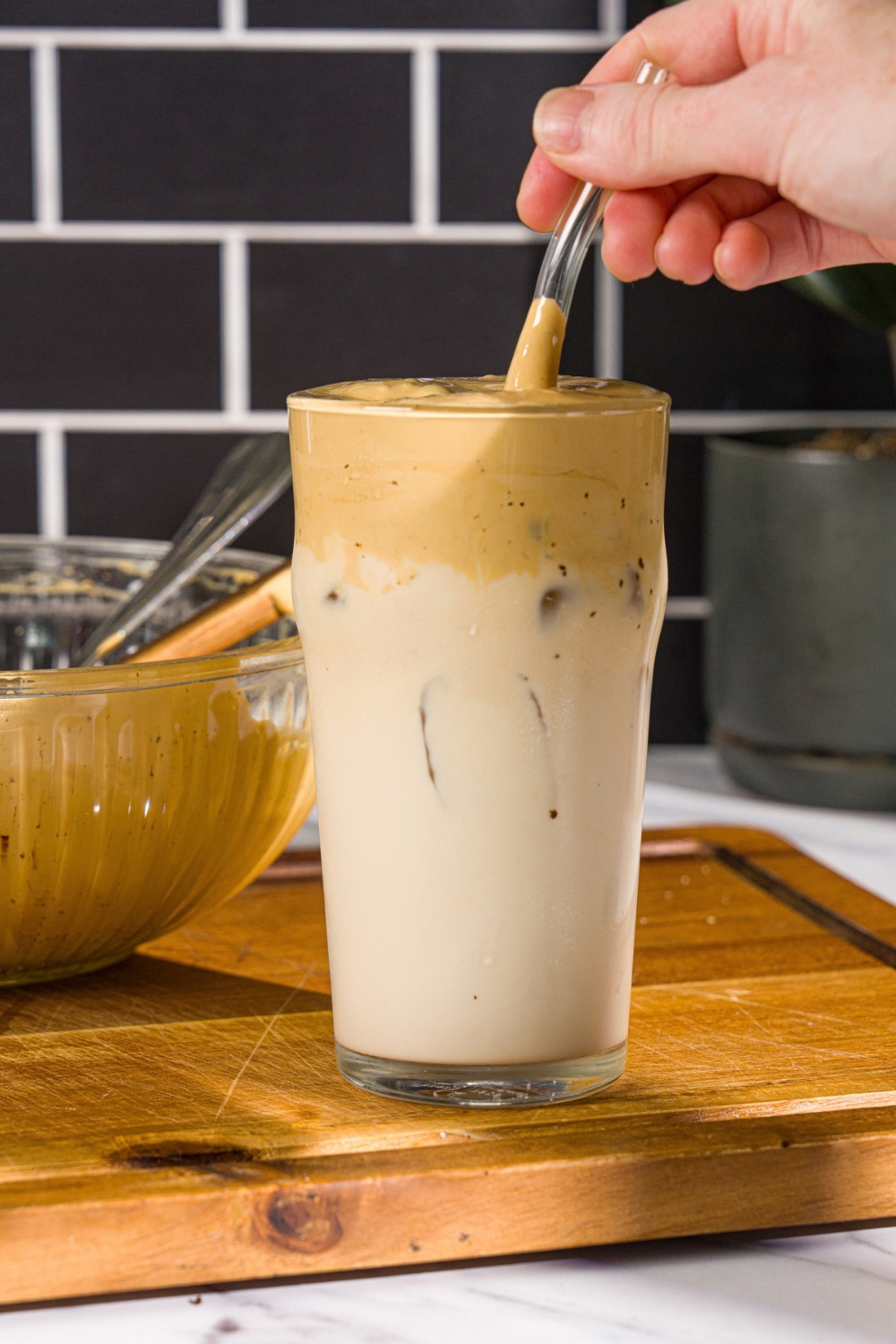 A glass with whipped honey iced coffee with milk. There is a spoon stirring the coffee in the glass. The glass is on a wooden board with a green cloth napkin.