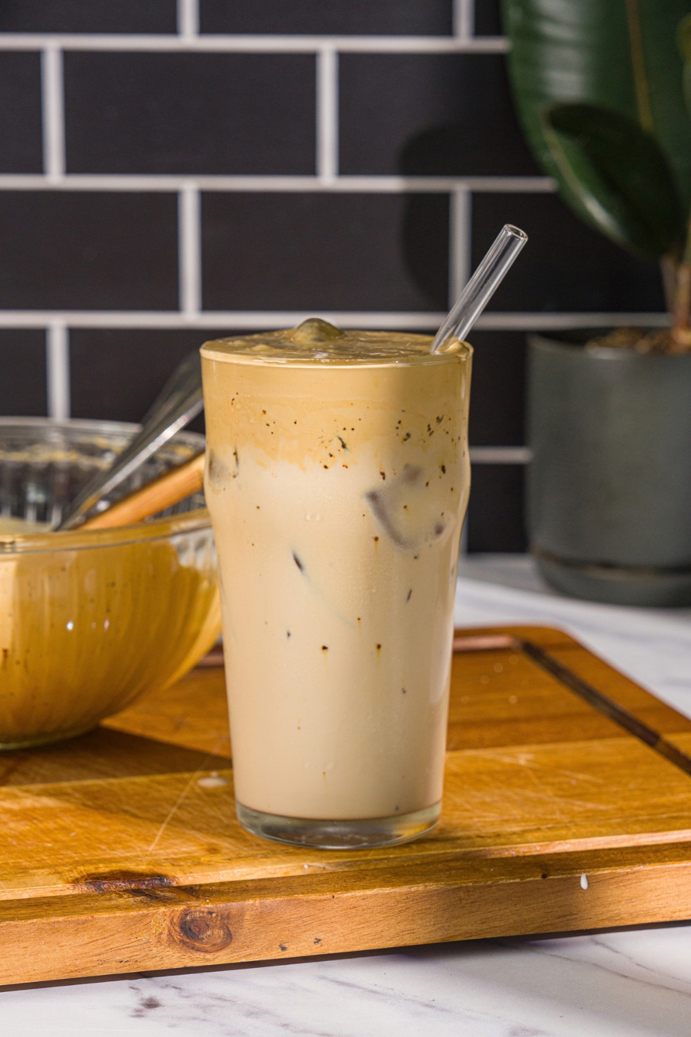 A glass with whipped honey iced coffee with milk. There is a spoon in the glass. The glass is on a wooden board with a bowl of whipped honey coffee.