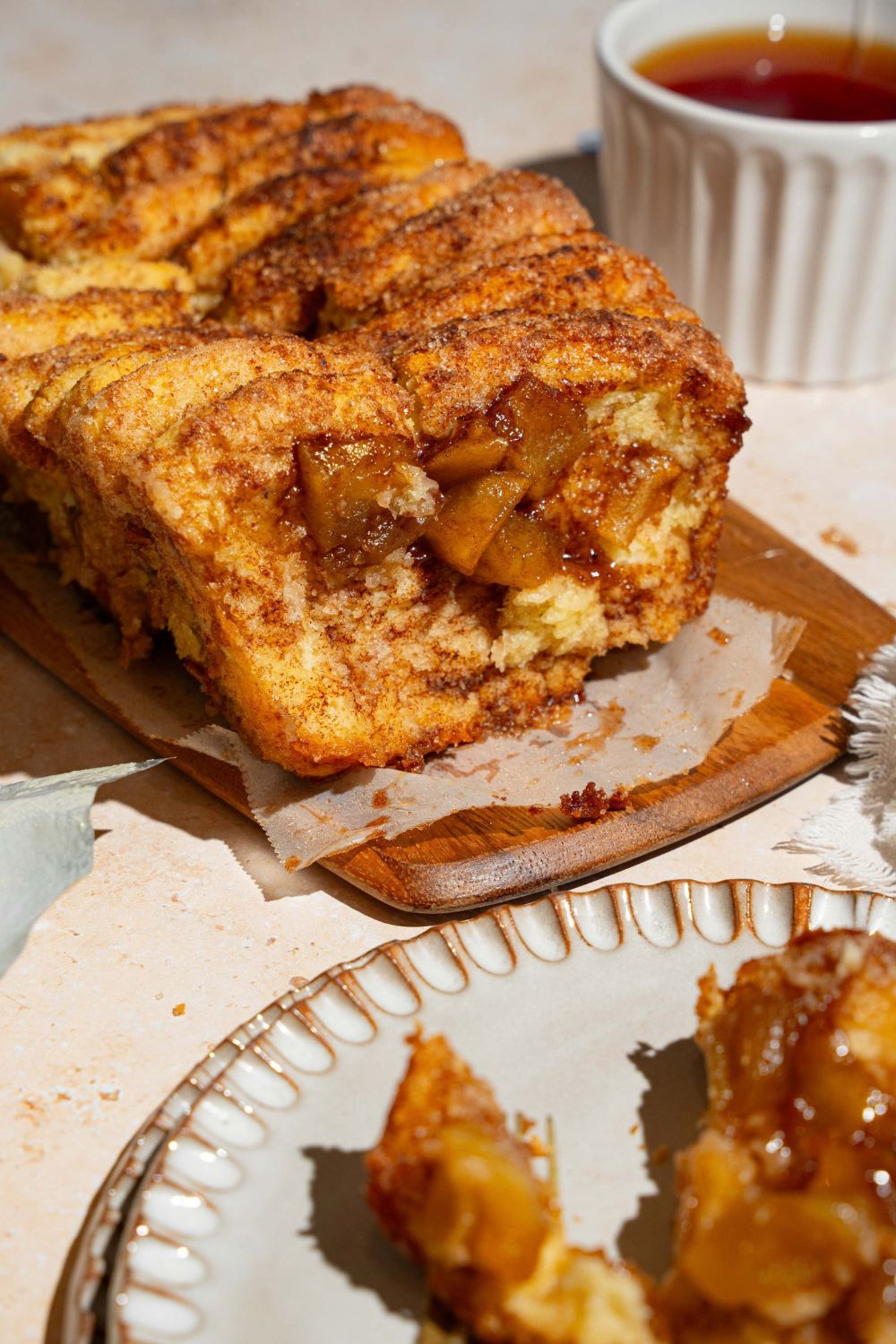 A loaf of apple pie pull apart bread on a wooden board lined with parchment paper. The board is on a tan counter with a plate with a slice of bread.