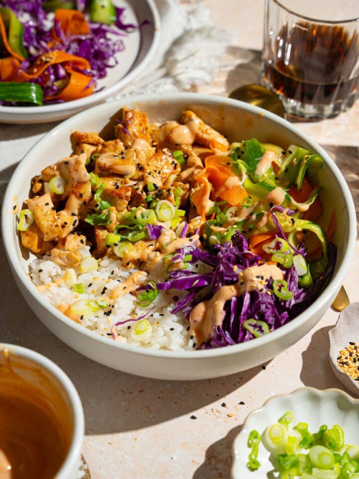 A bang bang chicken rice bowl topped with sliced green onion, cabbage, cucumber carrot salad, and sesame seeds. The bowl is on a tan counter with small bowls of toppings.