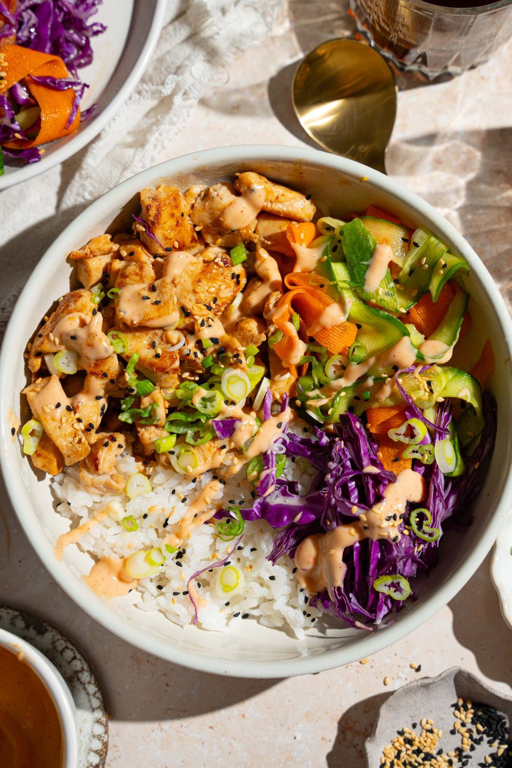 A bang bang chicken rice bowl topped with sliced green onion, cabbage, cucumber carrot salad, and sesame seeds. The bowl is on a tan counter with small bowls of toppings.