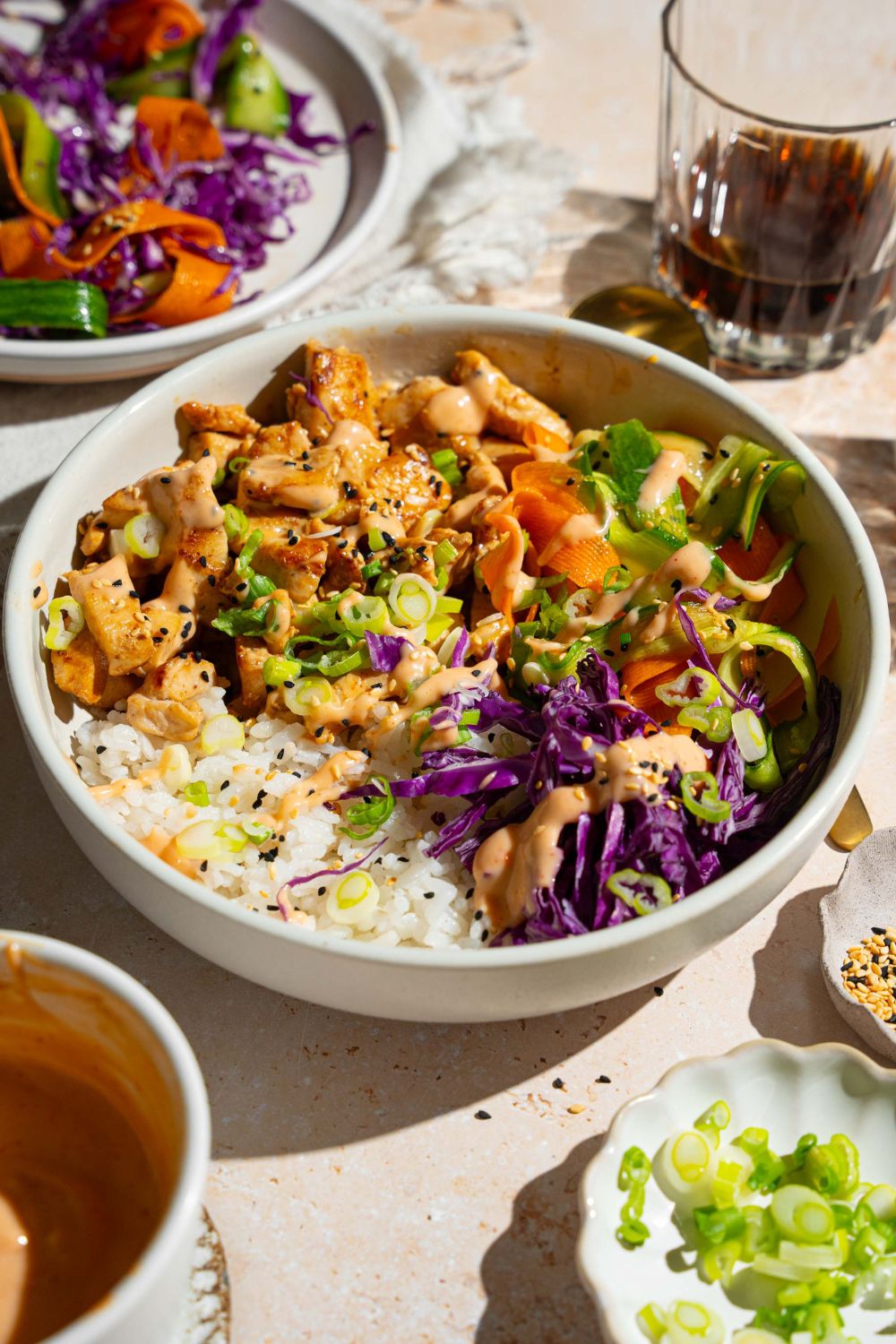 A bang bang chicken rice bowl topped with sliced green onion, cabbage, cucumber carrot salad, and sesame seeds. The bowl is on a tan counter with small bowls of toppings.