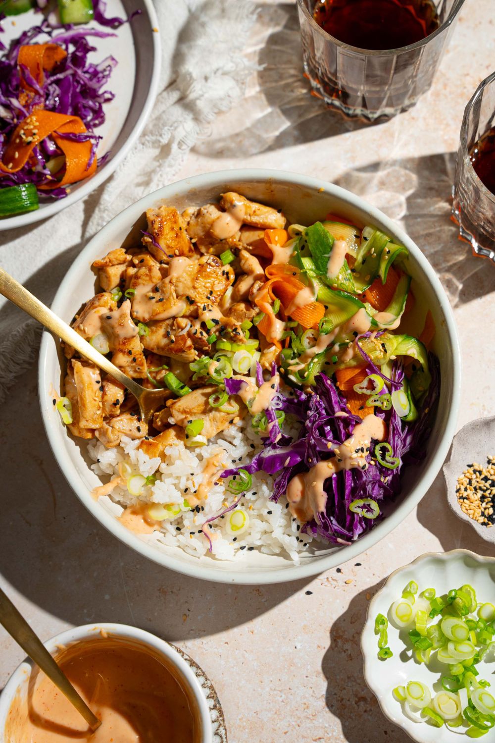 A bang bang chicken rice bowl topped with sliced green onion, cabbage, cucumber carrot salad, and sesame seeds. The bowl is on a tan counter with small bowls of toppings.