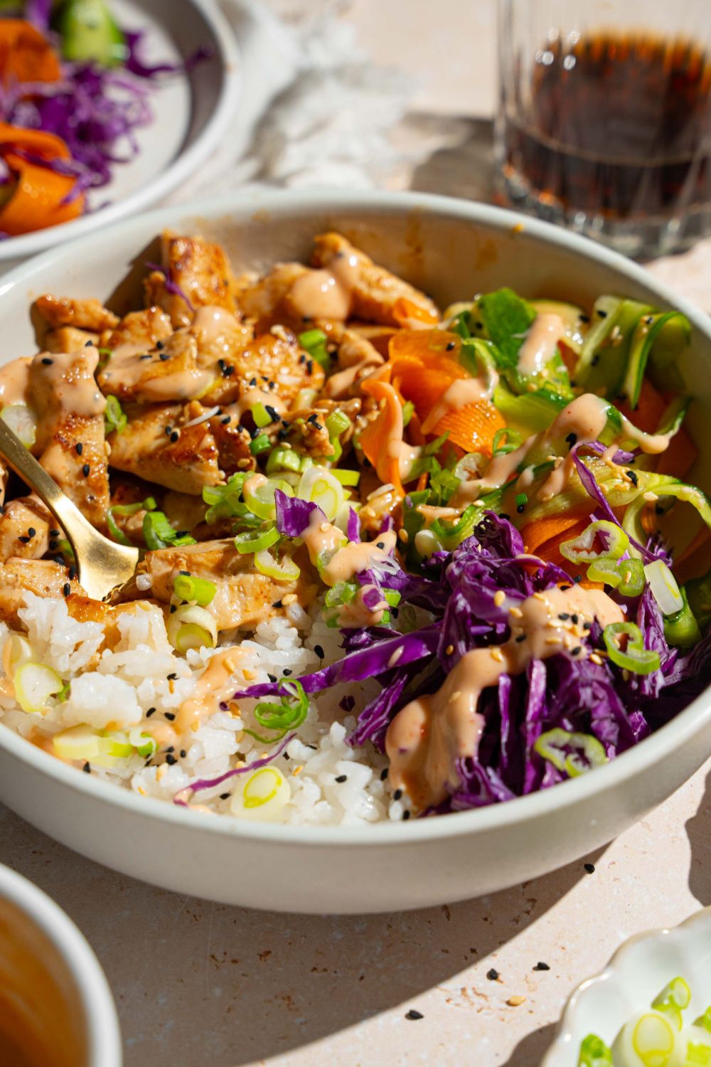 A bang bang chicken rice bowl topped with sliced green onion, cabbage, cucumber carrot salad, and sesame seeds. The bowl is on a tan counter with small bowls of toppings.