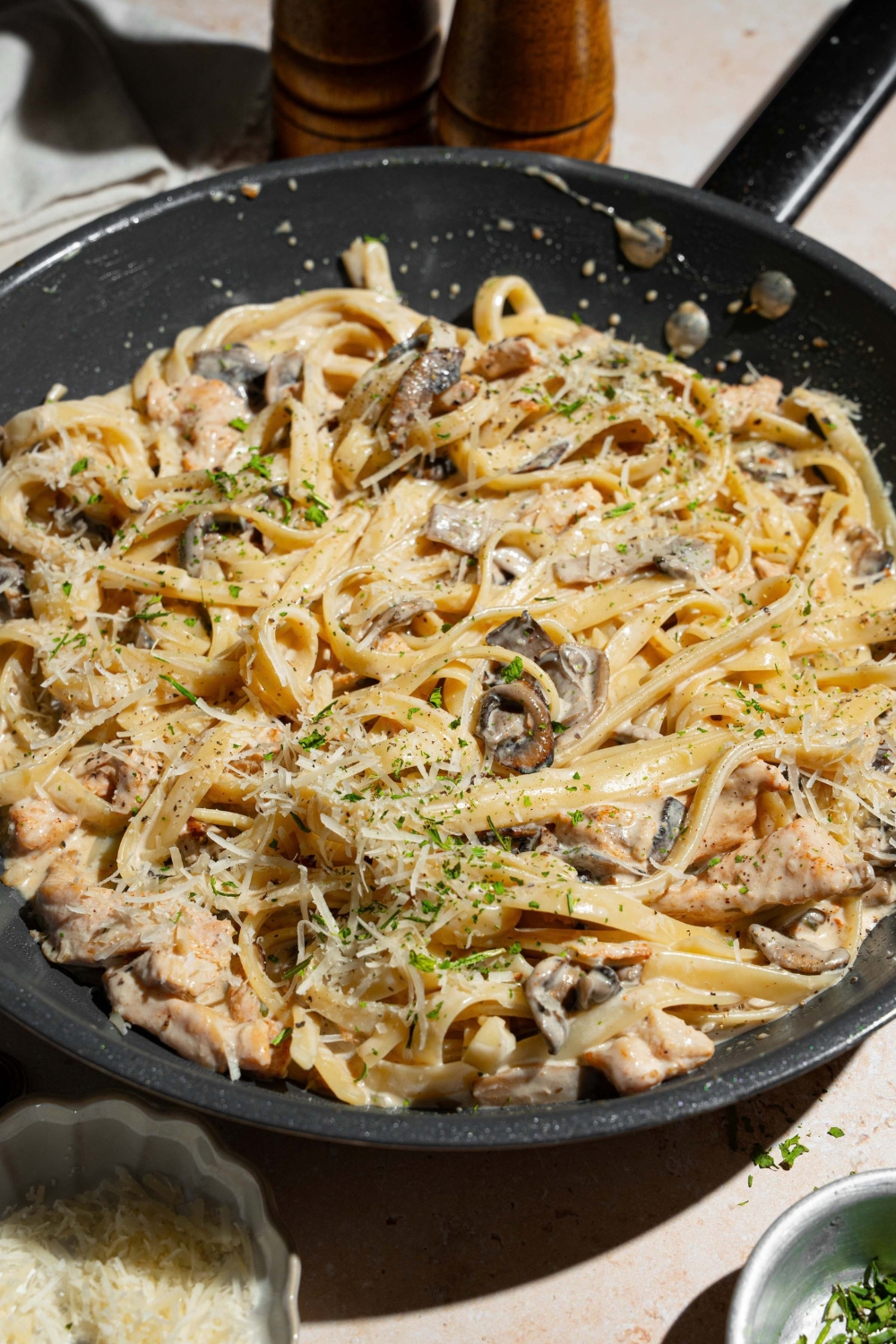 A skillet with chicken mushroom fettuccine alfredo garnished with pepper and parmesan. The skillet is on a tan counter with bowls of garnishes.