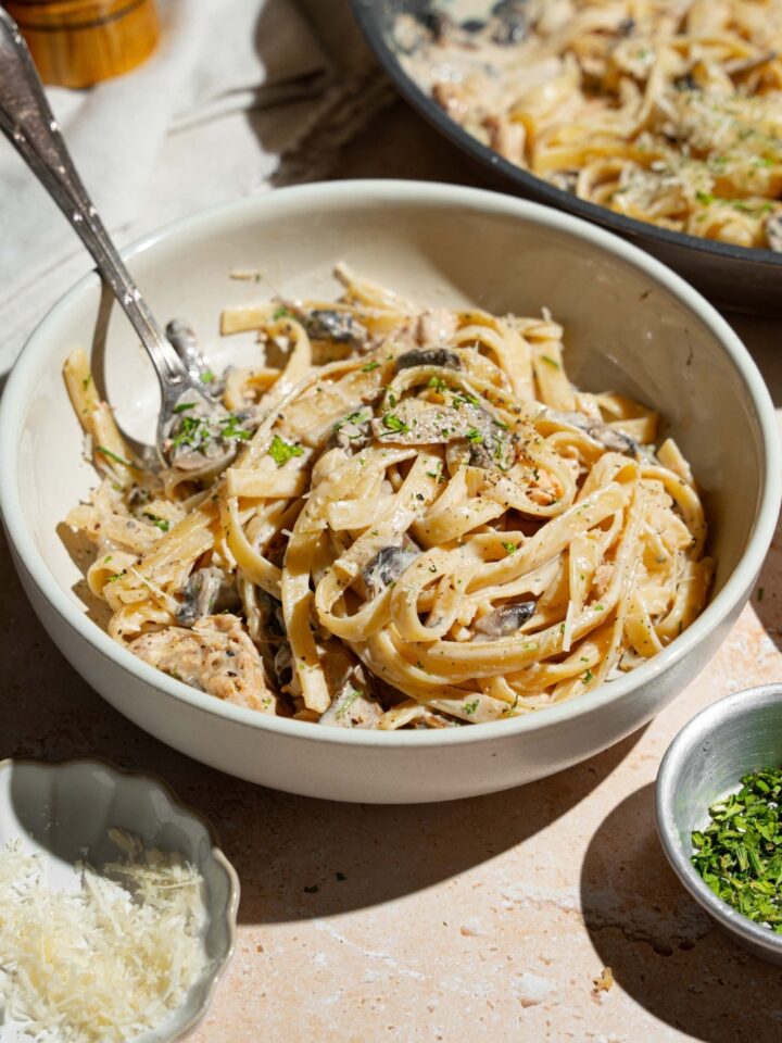 A white plate with chicken mushroom fettuccine alfredo garnished with pepper and parmesan. There is a fork in the pasta. The bowl is on a tan counter with small bowls of garnishes.