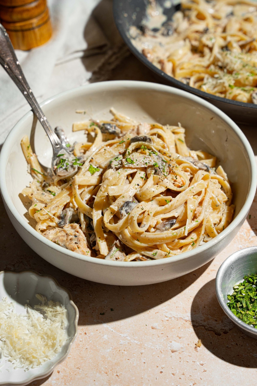 A white plate with chicken mushroom fettuccine alfredo garnished with pepper and parmesan. There is a fork in the pasta. The bowl is on a tan counter with small bowls of garnishes.