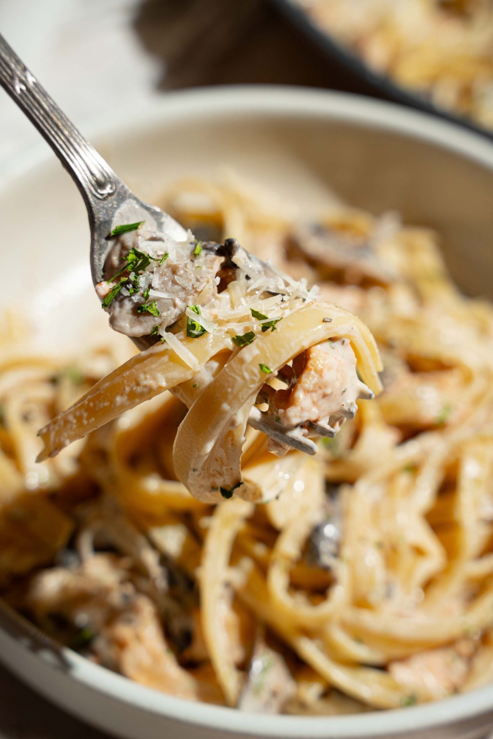 A close up of a fork with a bite of chicken mushroom fettuccine alfredo garnished with pepper and parmesan. There is a bowl of pasta blurred in the background.
