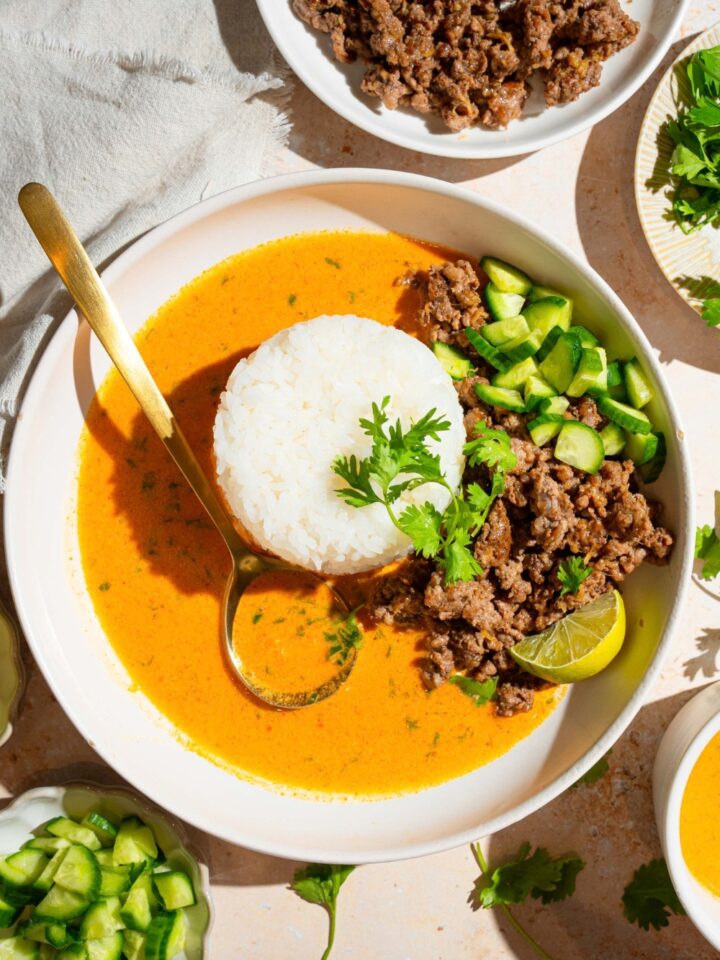 Ground beef, cucumbers, lime, wedge, and a pile of white rice in a coconut curry broth in a white bowl.