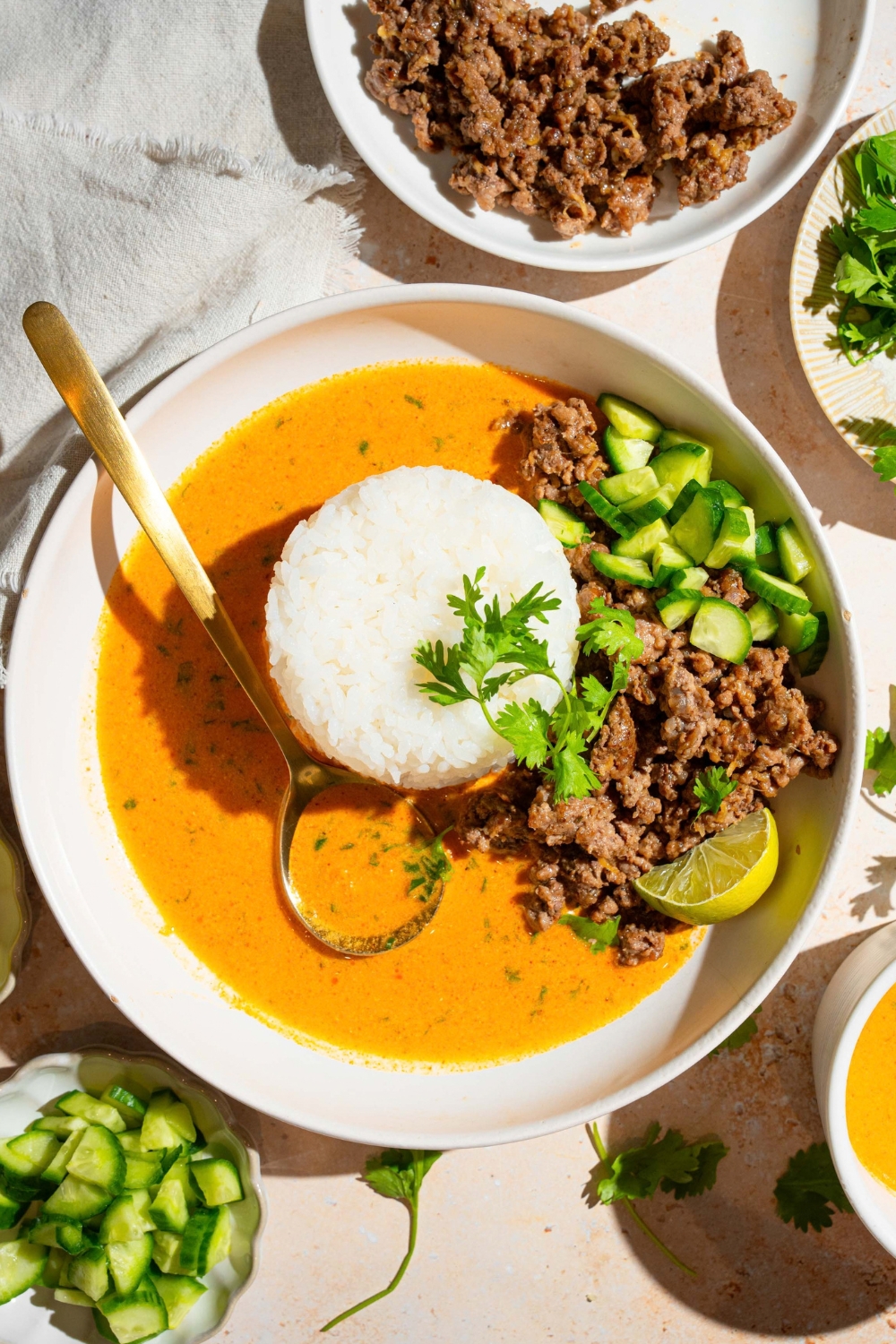 Ground beef, cucumbers, lime, wedge, and a pile of white rice in a coconut curry broth in a white bowl.