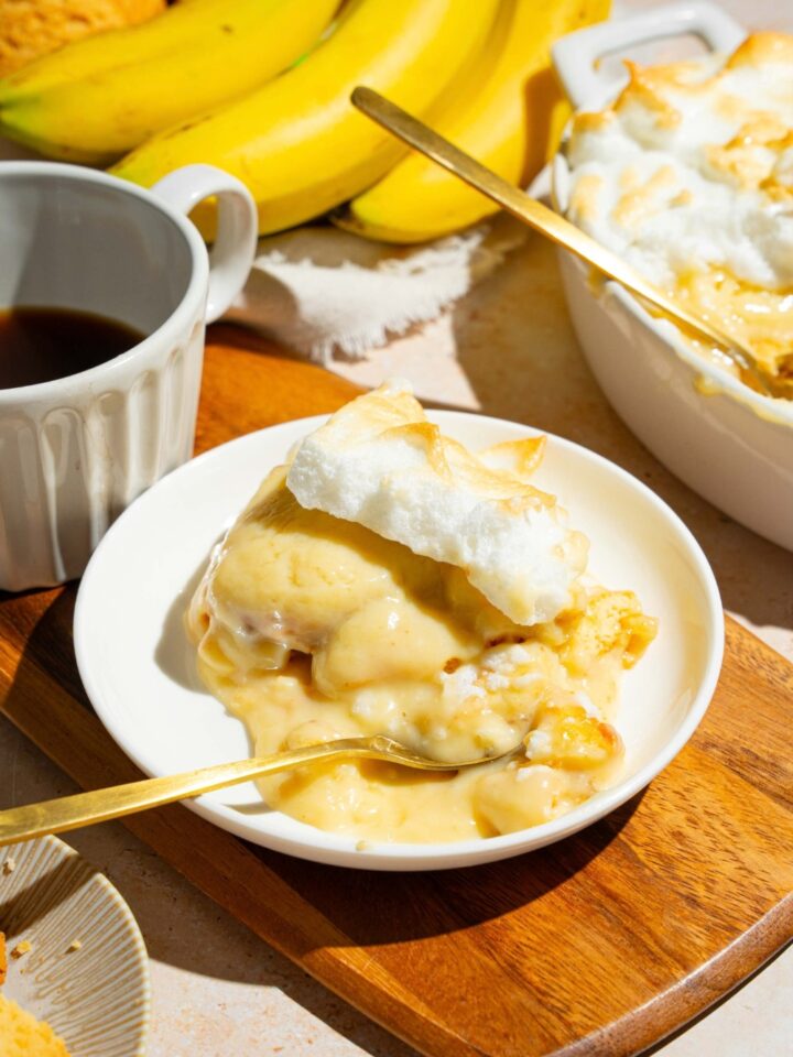 A slice of Dolly Parton's banana pudding on a white plate that is on a wood cutting board. Part of the banana pudding in a casserole dishes behind it.