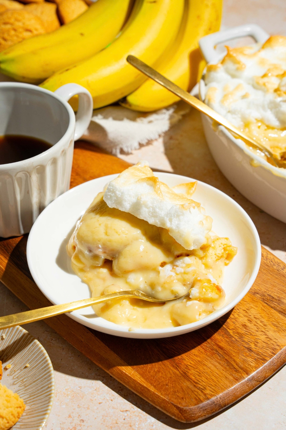 A slice of Dolly Parton's banana pudding on a white plate that is on a wood cutting board. Part of the banana pudding in a casserole dishes behind it.