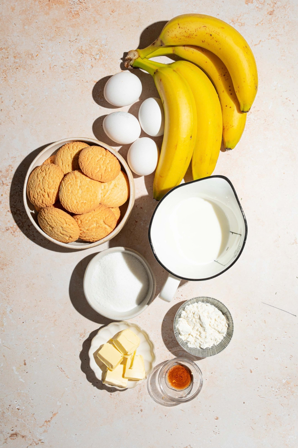Four eggs, four bananas, a bowl of vanilla wafer, cookies, a picture of milk, a bowl of sugar, bowl, flour, and a bowl of butter cubes all on a white counter.