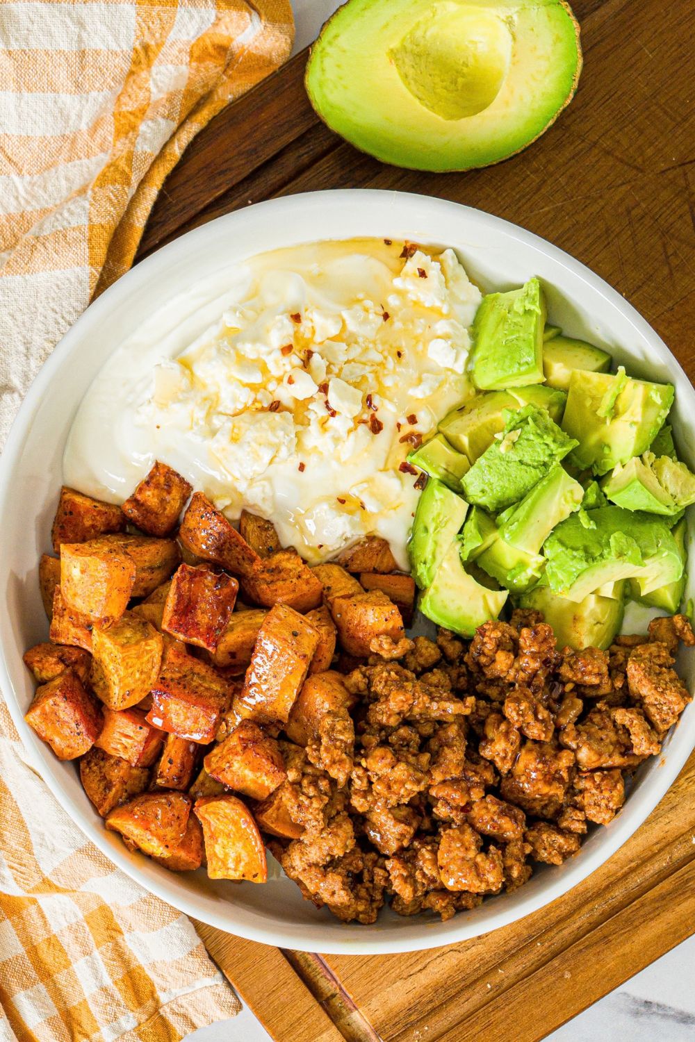 A white bowl with seasoned ground turkey, roasted sweet potatoes, avocado, and greek yogurt topped with hot honey. The bowl is on a wooden board with a yellow checkered napkin.