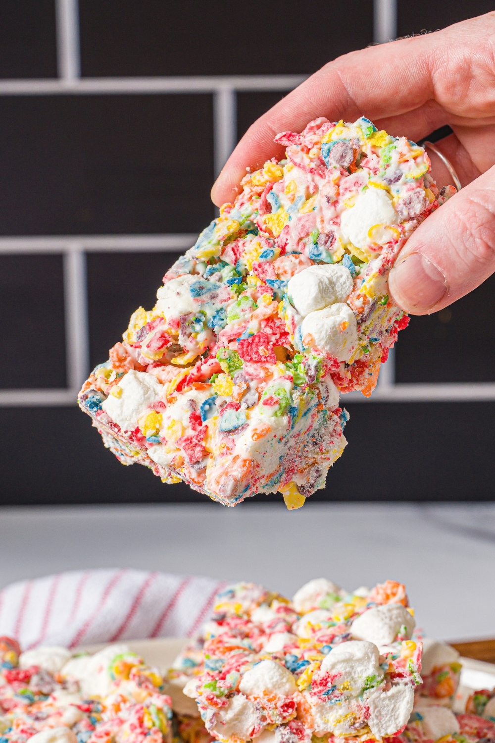 A hand holding a square of a fruity pebbles treat above a plate with a bunch of squares of the treat on it.