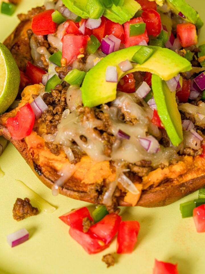 Avocado slices and diced red tomato and onion on top of melted cheese on ground beef that's all on a roasted sweet potato.