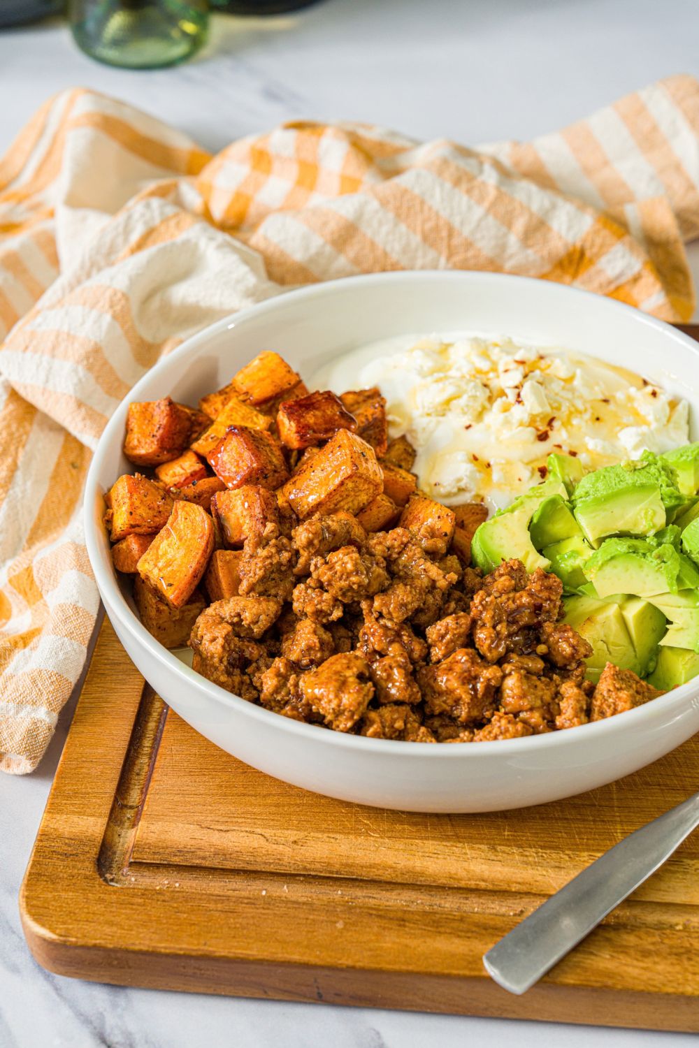 A white bowl with seasoned ground turkey, roasted sweet potatoes, avocado, and greek yogurt topped with hot honey. A fork is taking a bite from the bowl. The bowl is on a wooden board with a yellow checkered napkin.