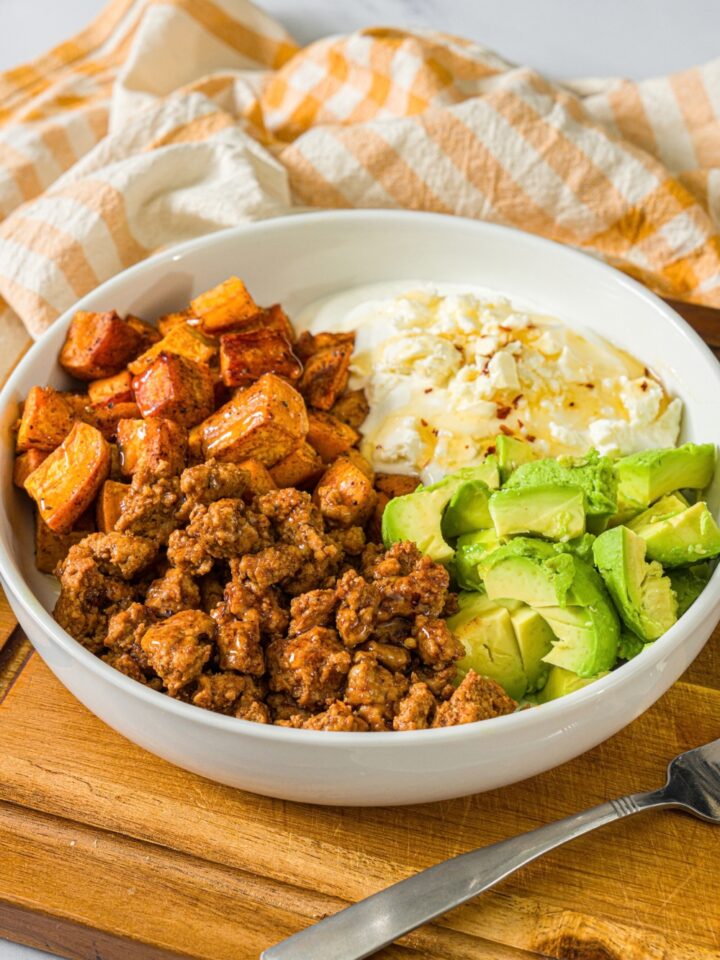 A white bowl with seasoned ground turkey, roasted sweet potatoes, avocado, and greek yogurt topped with hot honey. A fork is taking a bite from the bowl. The bowl is on a wooden board with a yellow checkered napkin.