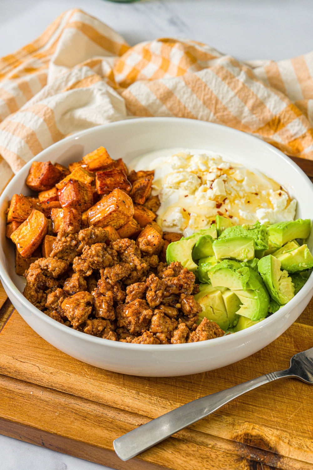A white bowl with seasoned ground turkey, roasted sweet potatoes, avocado, and greek yogurt topped with hot honey. A fork is taking a bite from the bowl. The bowl is on a wooden board with a yellow checkered napkin.