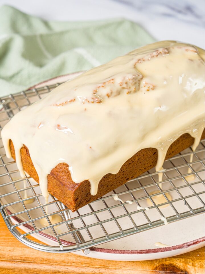 A white plate with a wire rack with a loaf of pumpkin bread with maple glaze. The plate is on a wooden board.