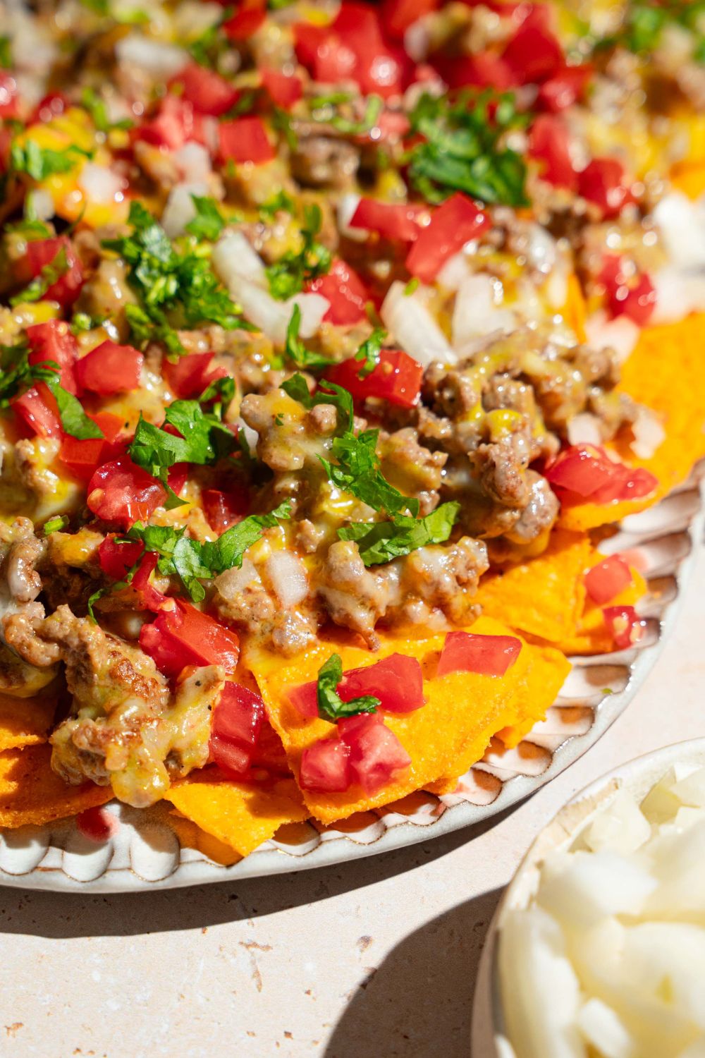 An oval platter with sliding sheet pan nachos topped with shredded cheese, ground beef, diced tomatoes and onions, and fresh parsley. The platter is on a tan counter.