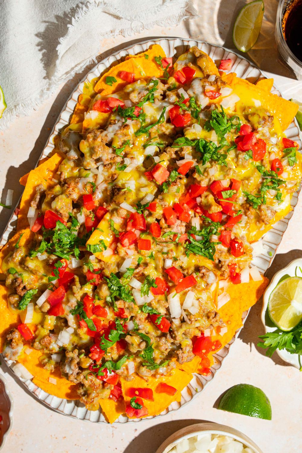 An oval platter with sliding sheet pan nachos topped with shredded cheese, ground beef, diced tomatoes and onions, and fresh parsley. The platter is on a tan counter with small plates of garnishes.