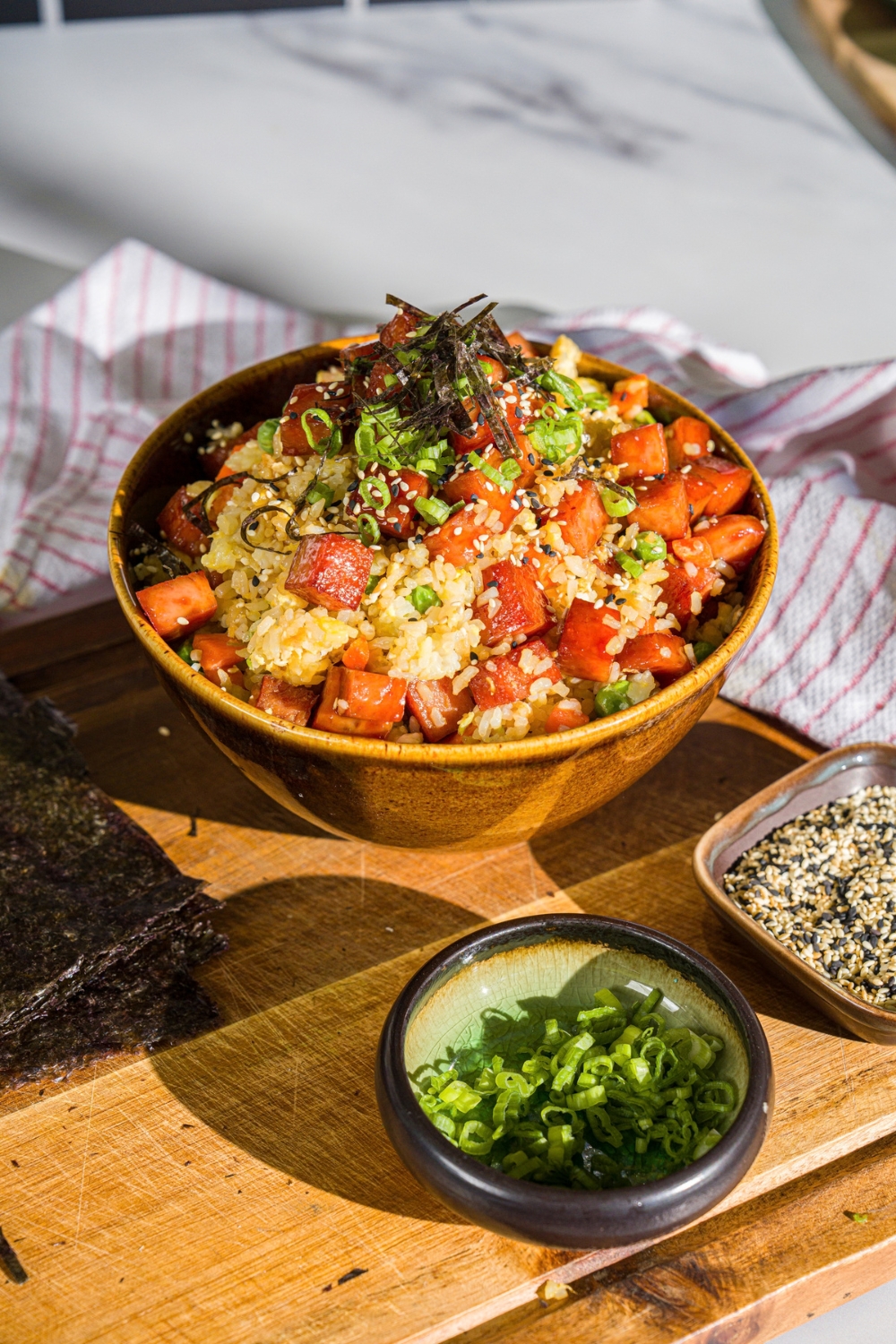 A spam musubi rice bowl garnished with seaweed, green onion, and sesame seeds. The bowl is on a wooden board with small bowls of garnishes.