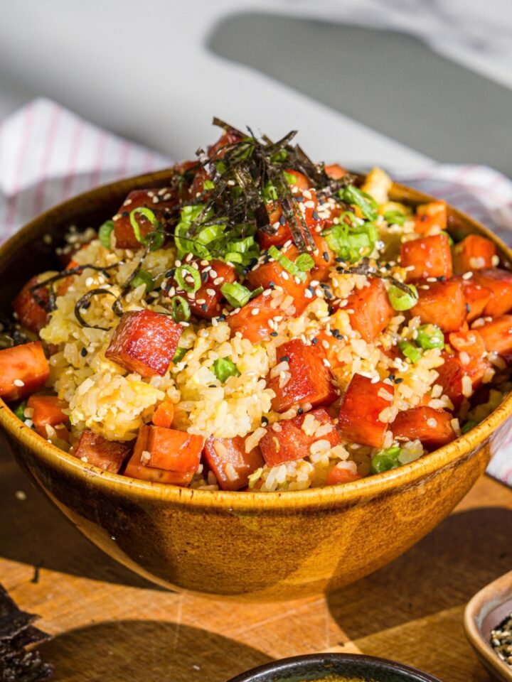 A spam musubi rice bowl garnished with seaweed, green onion, and sesame seeds. The bowl is on a wooden board with small bowls of garnishes.