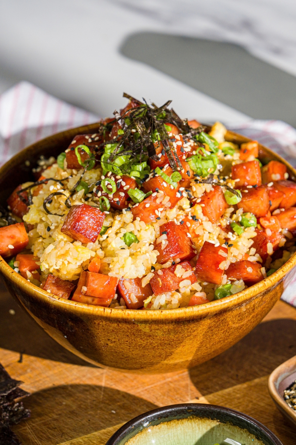 A spam musubi rice bowl garnished with seaweed, green onion, and sesame seeds. The bowl is on a wooden board with small bowls of garnishes.