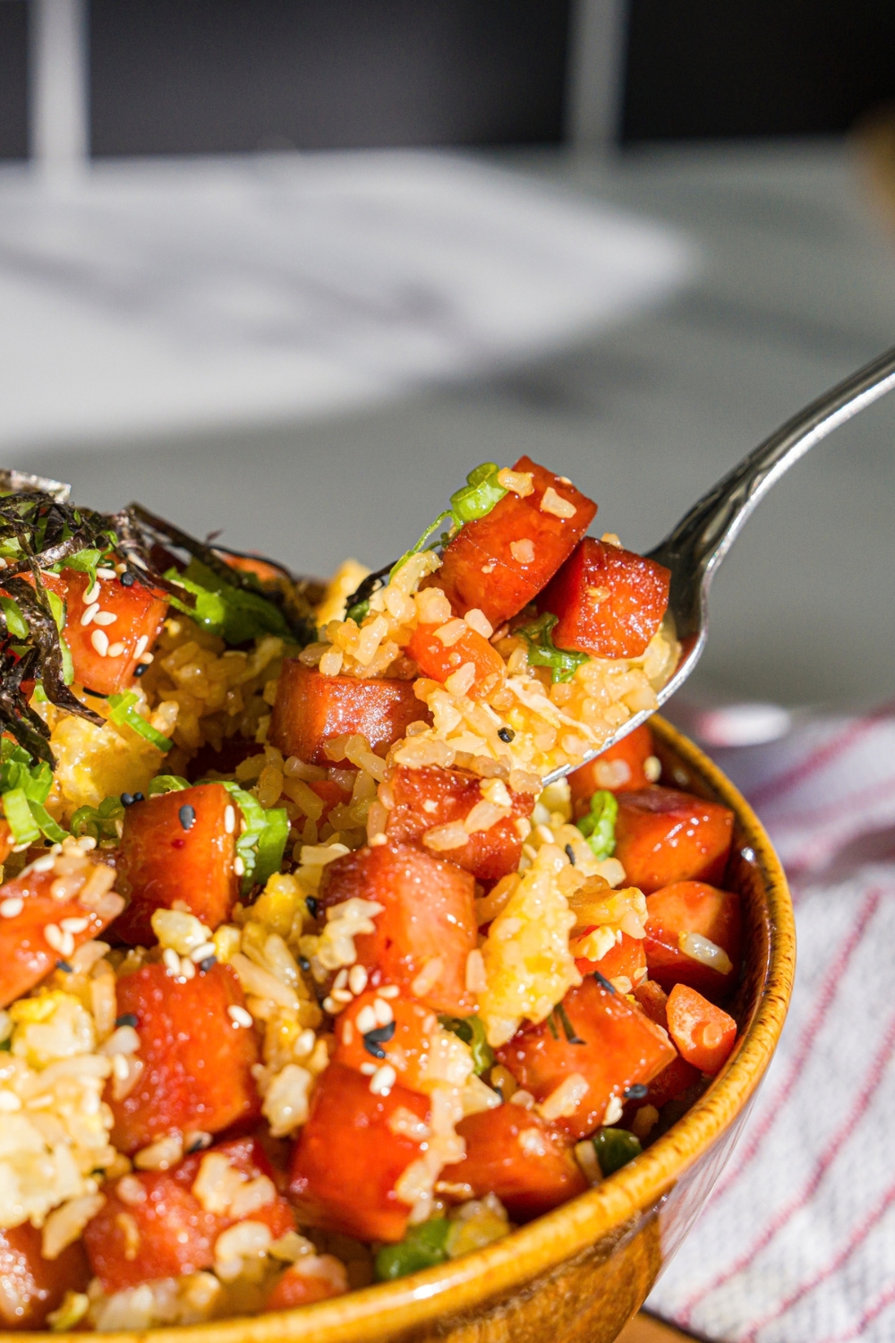 A spam musubi rice bowl garnished with seaweed, green onion, and sesame seeds. A fork is taking a bite from the bowl. The bowl is on a wooden board with small bowls of garnishes.