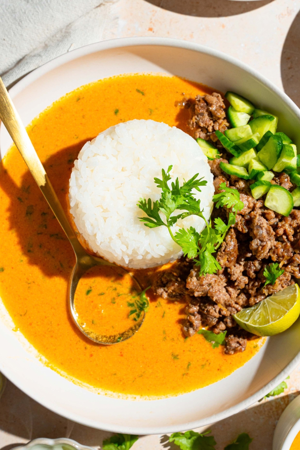 A bound of jasmine rice, ground beef, cucumber, and a lime wedge in a coconut curry broth, and a white bowl.