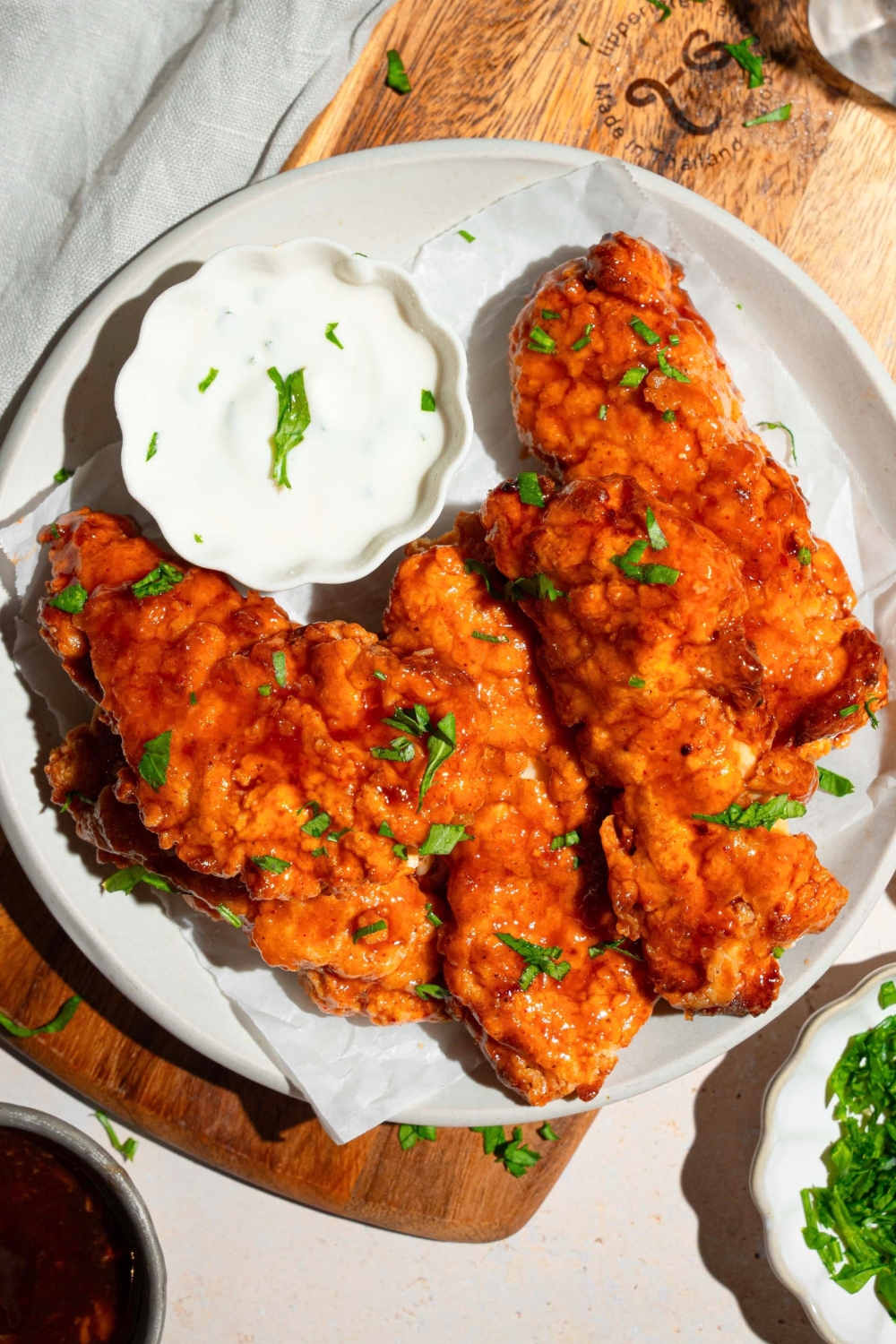 A white plate with sweet chili chicken tenders garnished with fresh parsley and served with a small bowl of dipping sauce. The plate is on a wooden board on a tan counter with garnishes.