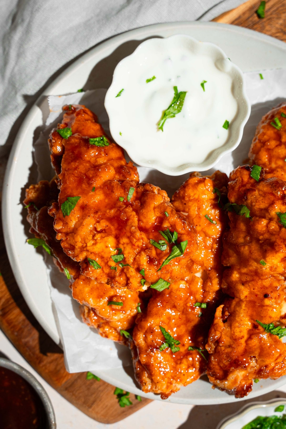 A white plate with sweet chili chicken tenders garnished with fresh parsley and served with a small bowl of dipping sauce. The plate is on a wooden board on a tan counter with garnishes.