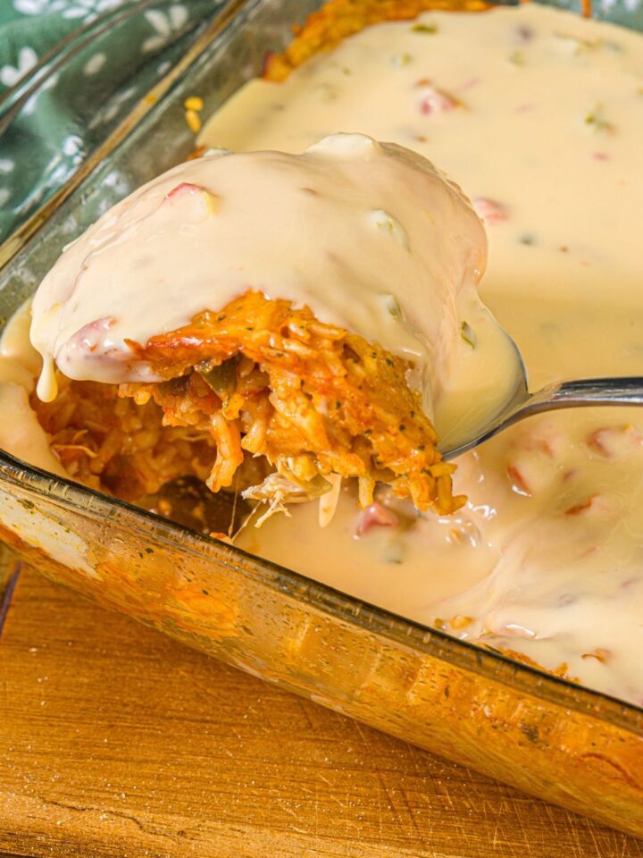 A baking dish with swim queso chicken and rice. A spoon is taking a scoop from the dish. The dish is on a wooden board.