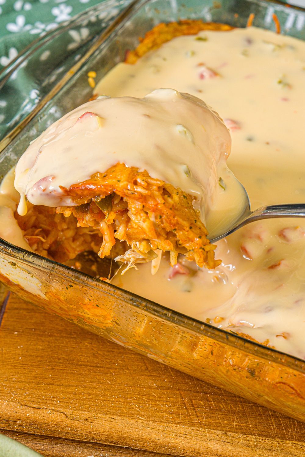A baking dish with swim queso chicken and rice. A spoon is taking a scoop from the dish. The dish is on a wooden board.