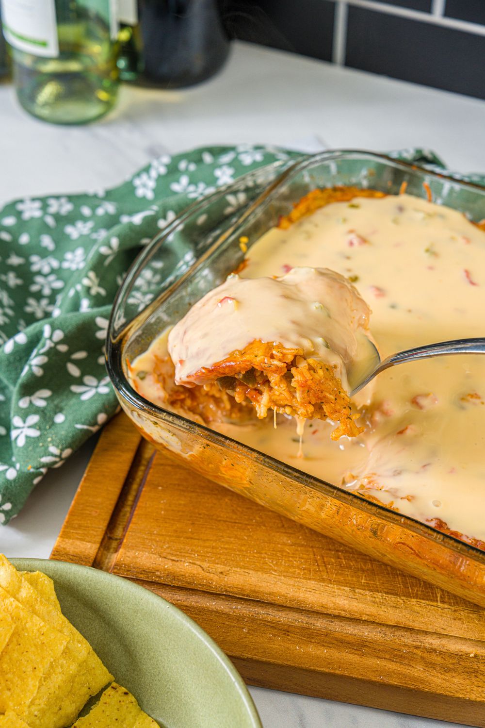 A baking dish with swim queso chicken and rice. A spoon is taking a scoop from the dish. The dish is on a wooden board with a green floral napkin.