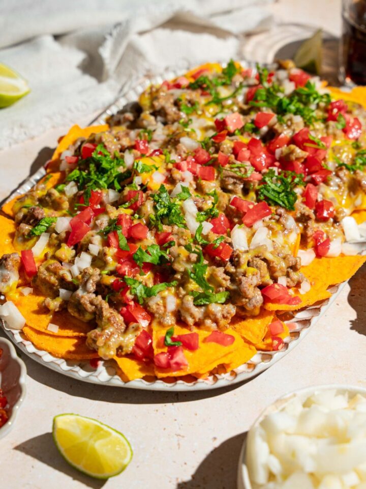 An oval platter with sliding sheet pan nachos topped with shredded cheese, ground beef, diced tomatoes and onions, and fresh parsley. The platter is on a tan counter with small garnishes.
