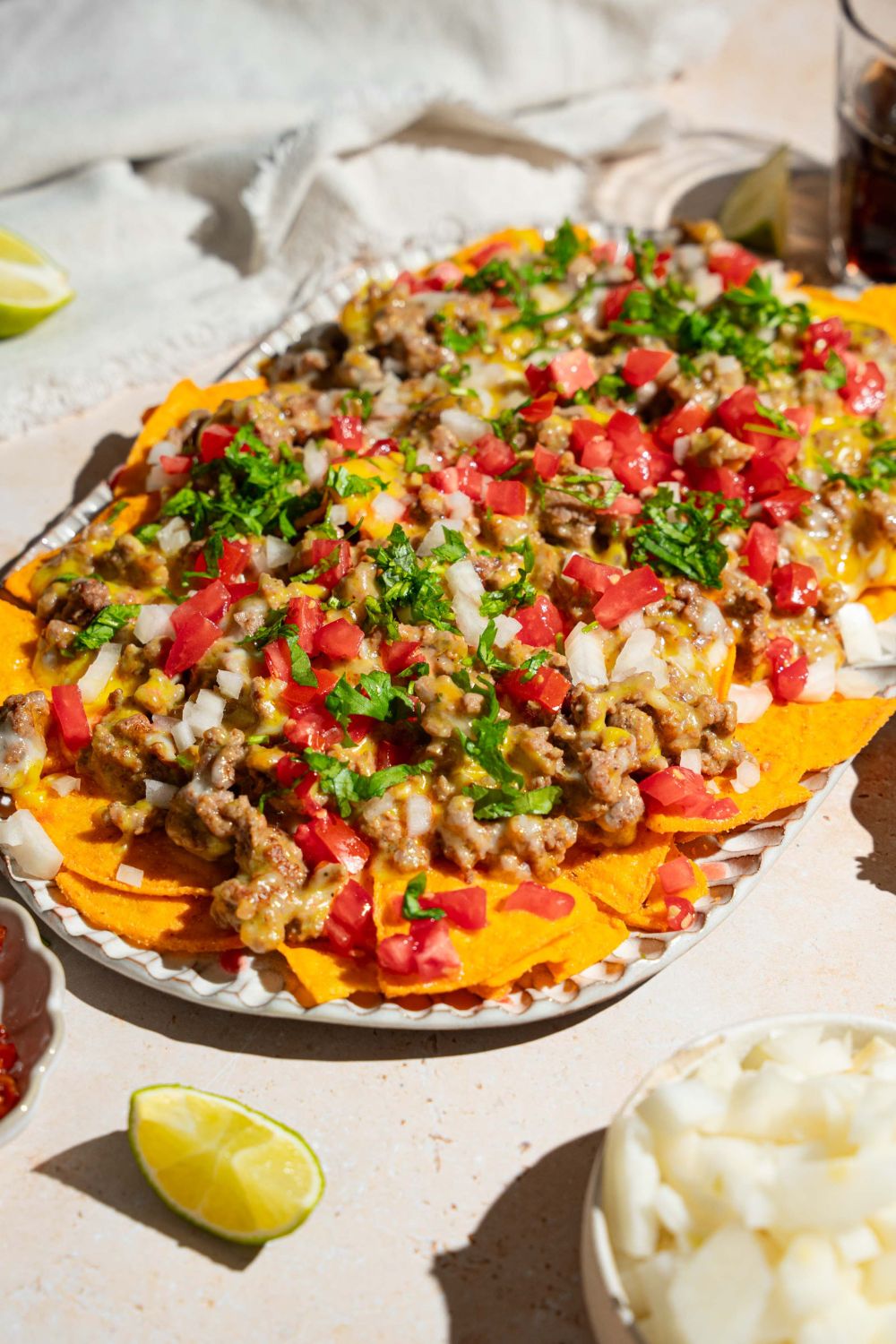 An oval platter with sliding sheet pan nachos topped with shredded cheese, ground beef, diced tomatoes and onions, and fresh parsley. The platter is on a tan counter with small garnishes.