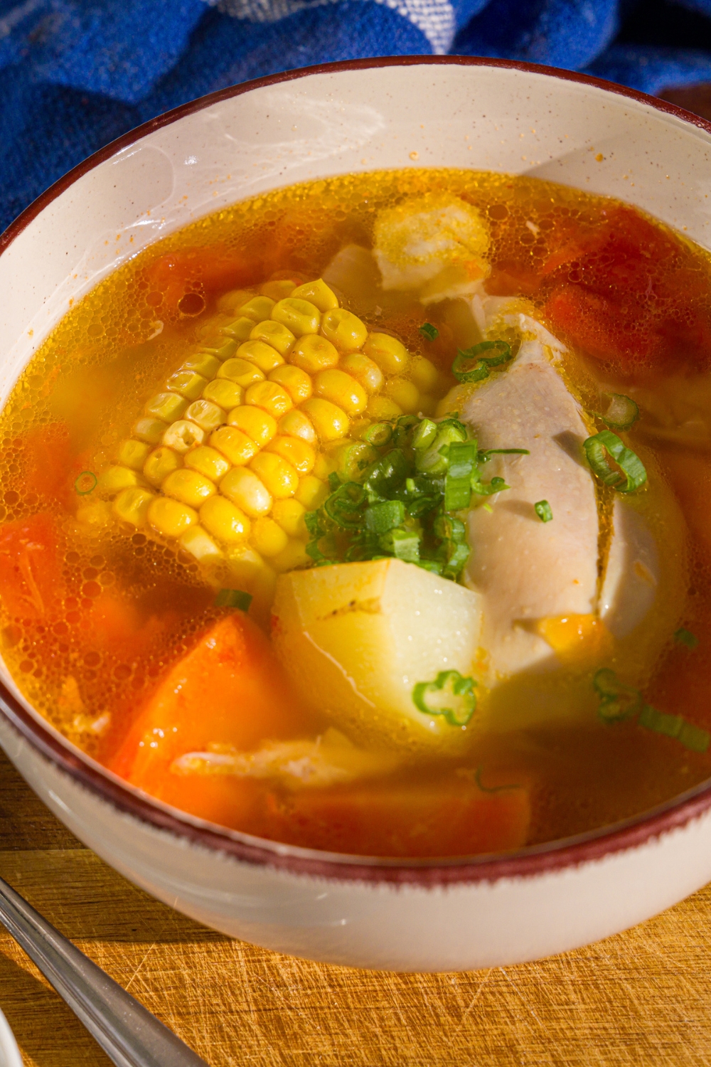 A bowl of ABC soup with corn on the cob, potatoes, carrots, and chicken garnished with sliced green onions. The bowl is on a wooden board with a spoon and a blue cloth napkin.