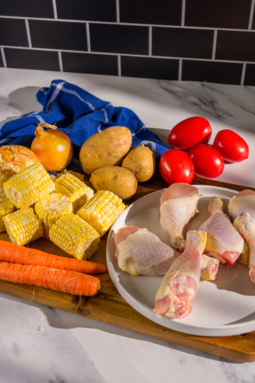 A wooden board with ingredients to make ABC soup including corn on the cob pieces, carrots, chicken drums, potatoes, onion, and tomatoes.