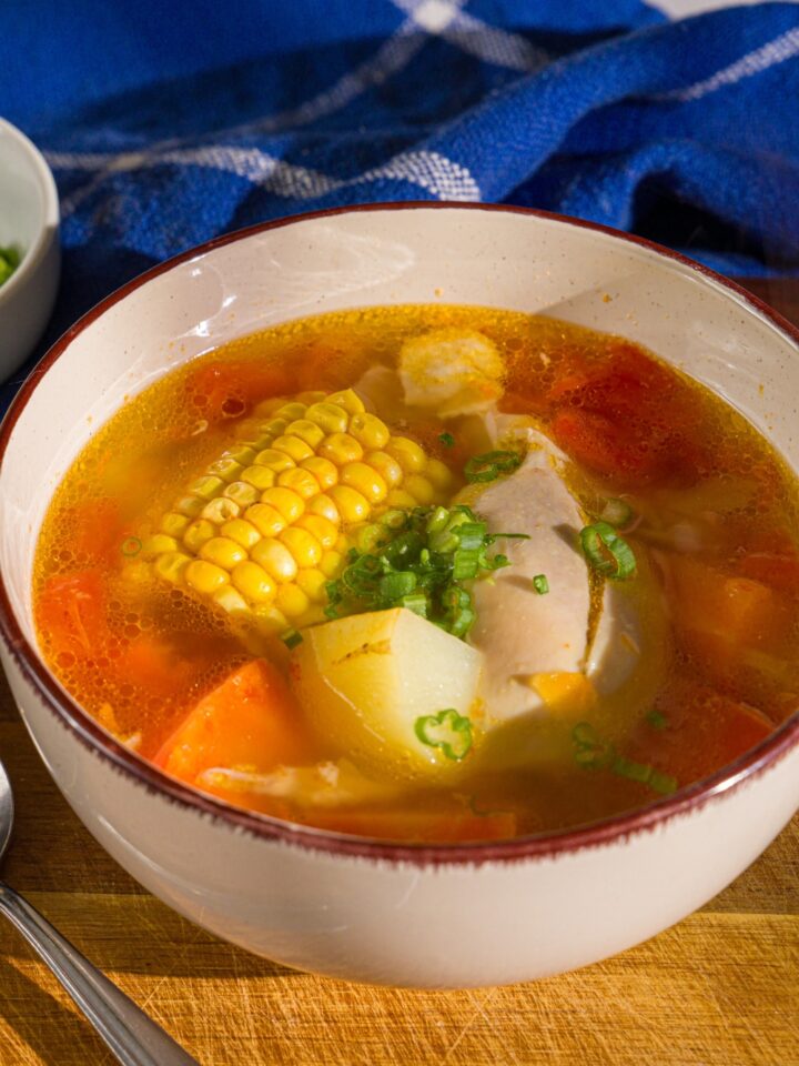 A bowl of ABC soup with corn on the cob, potatoes, carrots, and chicken garnished with sliced green onions. The bowl is on a wooden board with a spoon and a blue cloth napkin.