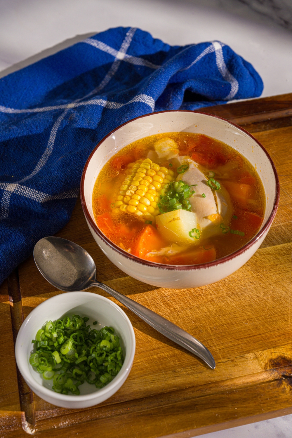 A bowl of ABC soup with corn on the cob, potatoes, carrots, and chicken garnished with sliced green onions. The bowl is on a wooden board with a spoon and a blue cloth napkin.