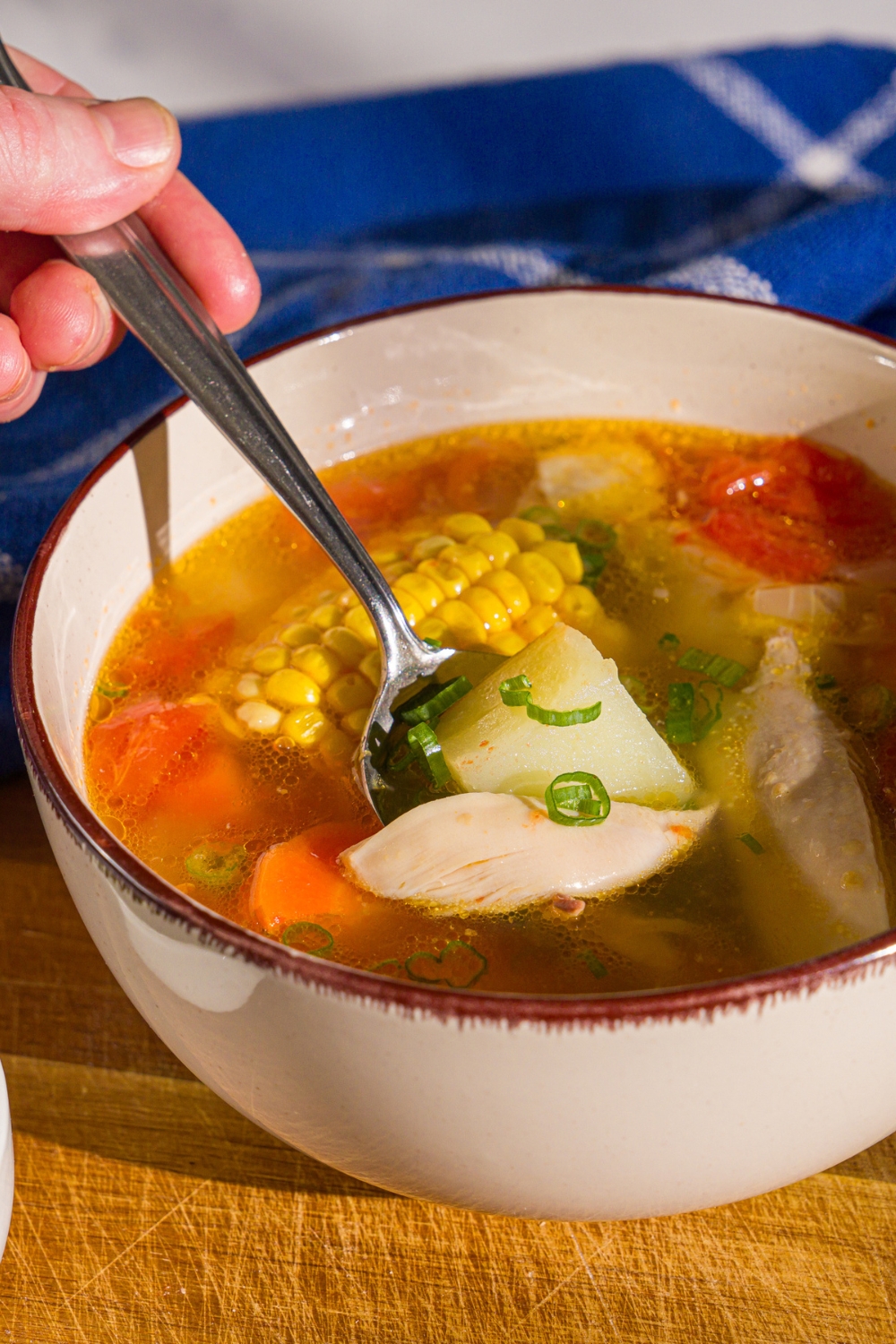A bowl of ABC soup with corn on the cob, potatoes, carrots, and chicken garnished with sliced green onions. The bowl is on a wooden board with a spoon dipped in the bowl.