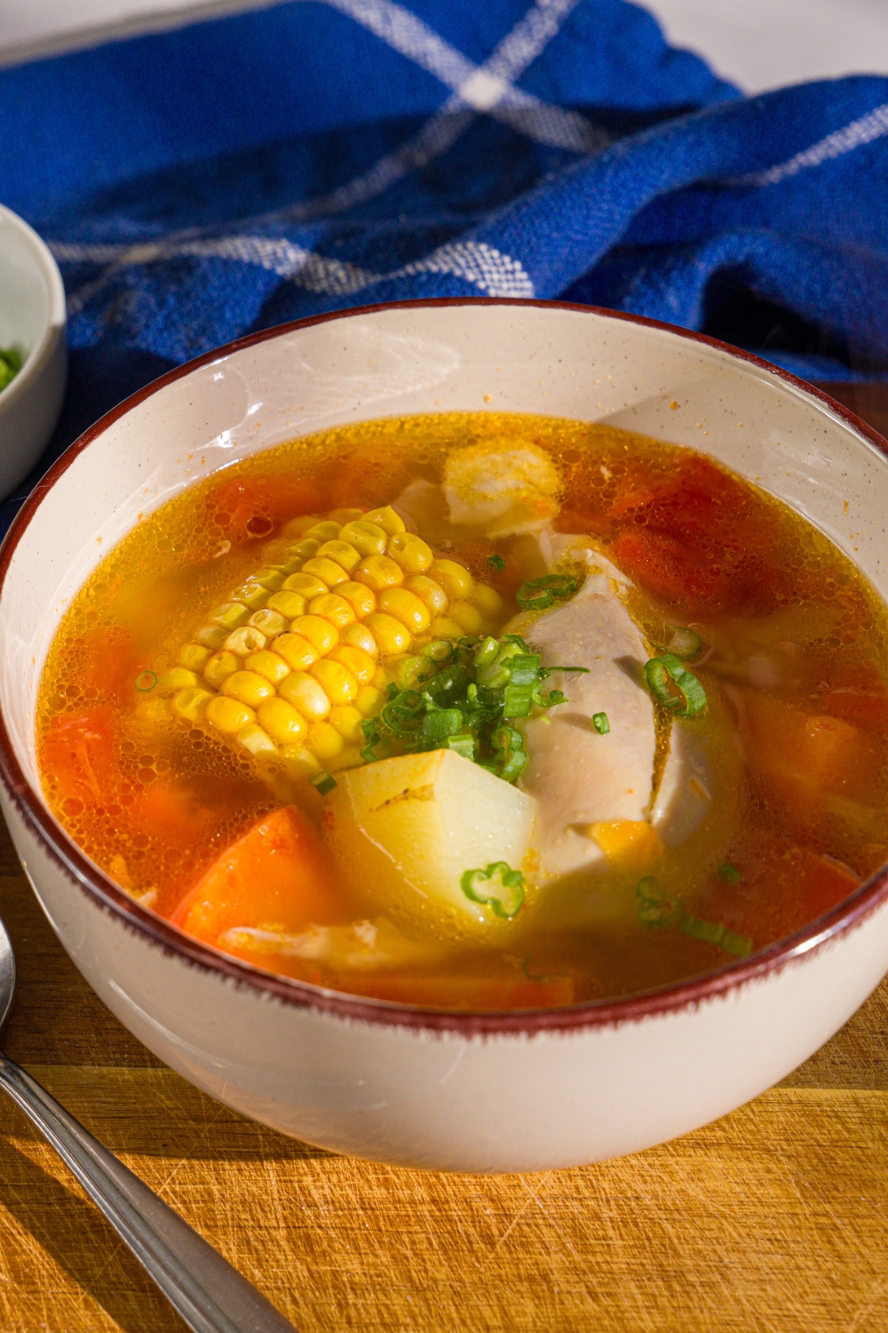 A bowl of ABC soup with corn on the cob, potatoes, carrots, and chicken garnished with sliced green onions. The bowl is on a wooden board with a spoon and a blue cloth napkin.